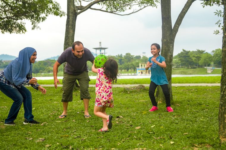 A family playing in a park, and little girl grab a ball and want to pass to her parent.