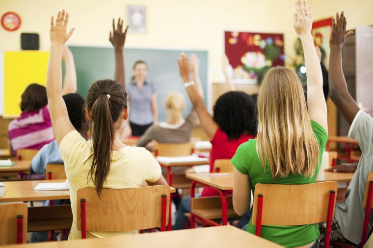 Rear view of the teens sitting in the classroom and raising hands to answer the question.