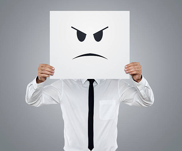 Young businessman holding card with an angry face on it isolated on gray background